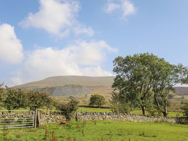 A rural landscape with a stone wall metal gate trees and a hill under a partly cloudy sky at Ffynnon Bach in Deiniolen