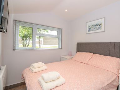 A bedroom with a bed covered in pink bedding and folded towels on top a window with blinds a wall-mounted television a nightstand with a lamp and a framed picture on the wall at 201 Norton Park in Dartmouth