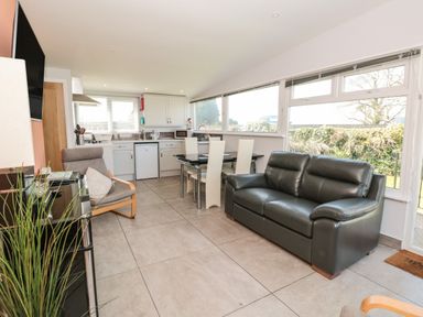 An open living area with a black leather sofa, a dining table with white chairs, a grey armchair, kitchen cabinets, and large windows at 201 Norton Park in Dartmouth
