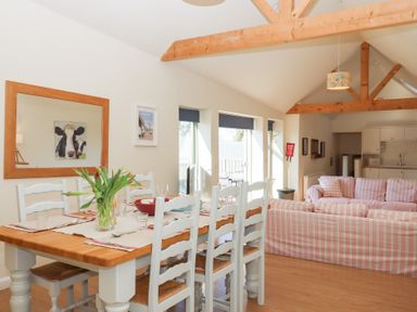 A dining area with a wooden table and white chairs next to a living room with pink striped sofas and exposed wooden beams at The Shippon at Wyke Farm in Lyme Regis