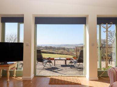 A room with large open doors leading to a patio with two chairs and a table overlooking a rural landscape at The Shippon at Wyke Farm in Lyme Regis