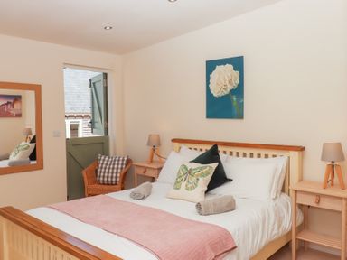 A bedroom with a bed with pillows and towels a wicker chair a mirror and bedside tables with lamps at The Shippon at Wyke Farm in Lyme Regis
