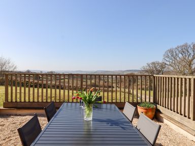 An outdoor dining table with a vase of flowers on a terrace overlooking fields and hills at The Shippon at Wyke Farm in Lyme Regis