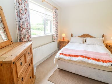 A bedroom with a double bed two bedside tables with lamps a wooden dresser with mirror and a window with floral curtains at Sea View Hideaway in Lahinch County Clare
