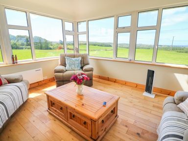 A sitting room with armchairs and a wooden coffee table with flowers by windows showing green fields at Sea View Hideaway in Lahinch County Clare