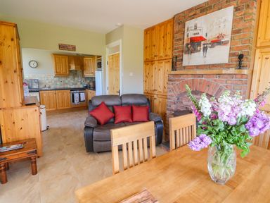 A dining area with a vase of flowers on the table a gray loveseat with red pillows wooden cabinets a brick fireplace and a kitchen with wooden cabinets in the background at Sea View Hideaway in Lahinch County Clare