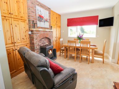 A dining area with a wooden table and chairs next to a window with a red blind and a seating area with grey recliners and a brick fireplace at Sea View Hideaway in Lahinch County Clare
