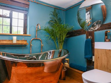 A bathroom with a copper bath tub blue walls a round mirror green plant and wooden accents at 3 Masons Court Stratford-Upon-Avon