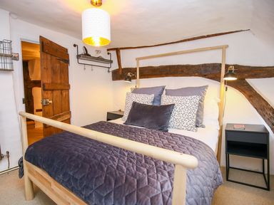 A bedroom with a wooden bed frame a quilted blanket pillows bedside tables wall lamps a wooden door and exposed wooden beams at 3 Masons Court in Stratford-Upon-Avon
