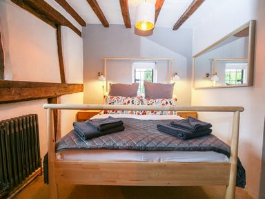 A bedroom with a double bed, wooden beams on ceiling and walls, bedside tables, lamps, and a rectangular mirror at 3 Masons Court Stratford-Upon-Avon