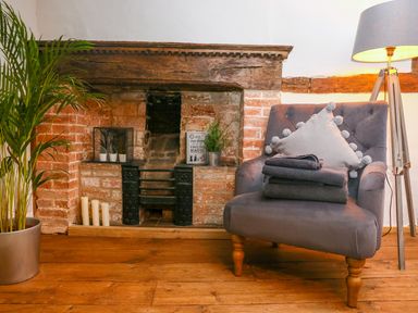 A grey armchair with folded towels and a cushion next to a brick fireplace with candles plant and a book at 3 Masons Court in Stratford-Upon-Avon