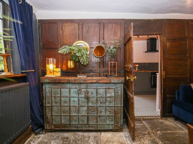 A wooden paneled room with an antique chest, decorative plants, a lamp, and an open door leading to a kitchen at 3 Masons Court in Stratford-Upon-Avon