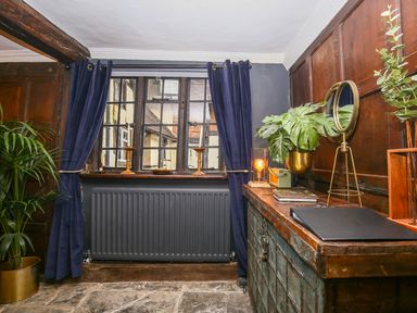 A room with a window and blue curtains a radiator a wooden cabinet with plants candles a lamp and a mirror at 3 Masons Court Stratford-Upon-Avon