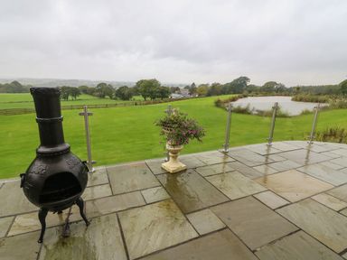 A patio with a chiminea and a planter with flowers overlooking a grassy field and a pond at Nicky Nook Lodge in Scorton