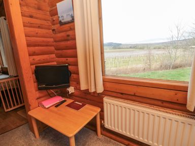 A corner of a wooden room with a small table holding a remote and books below a wall-mounted screen and a window with a view of fields at Glanton in Longframlington
