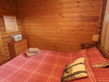 A bedroom with a wooden bed, red plaid bedspread, two towels on the bed, wooden dresser with a mirror, and a bedside table with a lamp in Glanton Longframlington