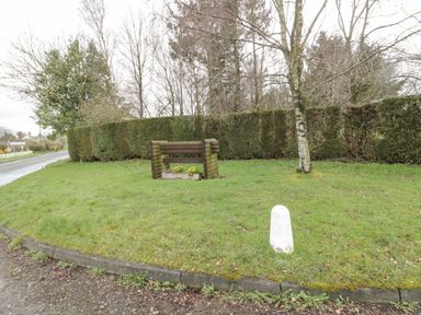 A grassy roadside area with a wooden sign and a tree near a hedge at Glanton in Longframlington