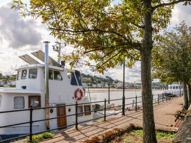 A docked boat next to a paved walkway with trees and benches along a river at The Shippon in Barnstaple