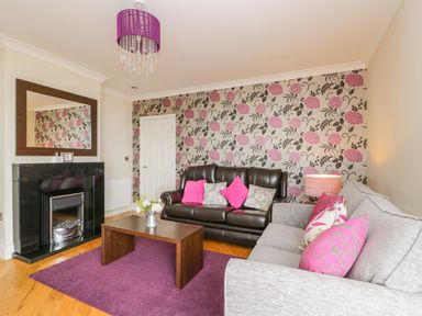 A living room with a dark brown sofa, a light gray sofa, a wooden coffee table, floral wallpaper, a purple rug, and a fireplace at The Gansey Girl in Bridlington