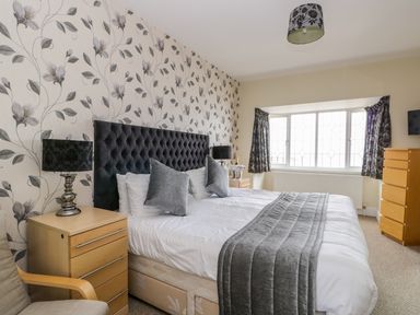 A bedroom with a large bed gray pillows and gray throw blanket beside wooden nightstands with lamps a window with curtains and a wooden chest of drawers at The Gansey Girl in Bridlington