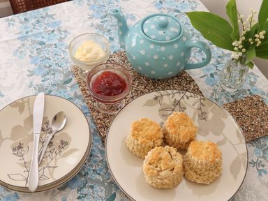 A table set with four scones on a plate a blue polka dot teapot jam cream in glass dishes utensils on plates and a vase with flowers on a floral tablecloth at Tremethic in Trewoon