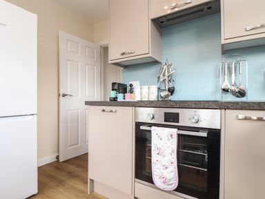 A kitchen with beige cabinets a black oven with a floral oven mitt on the handle kitchen utensils on the counter a white refrigerator and a door open at Tremethic in Trewoon