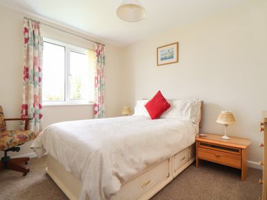 A bedroom with a bed with a red cushion bedside tables with lamps a window with floral curtains and a chair at Tremethic in Trewoon