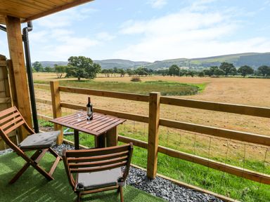 A wooden balcony with two chairs a table with a bottle and two glasses and a view of a field and hills at Cilan Lodge in Llandrillo