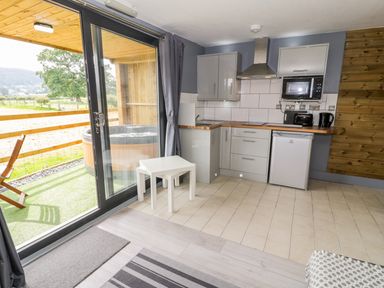 A small kitchen with gray cabinets microwave refrigerator and two white nested tables near sliding glass doors leading to an outdoor area with a hot tub and wooden chairs at Cilan Lodge in Llandrillo