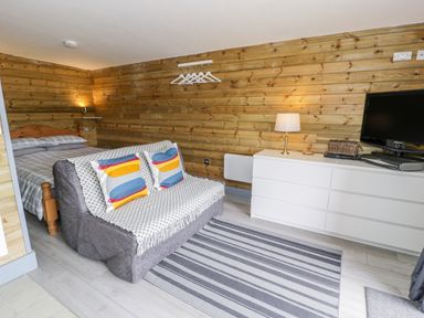 A bedroom with wooden walls containing a bed a small sofa with colorful pillows a striped rug a white dresser with a lamp and a TV at Cilan Lodge in Llandrillo