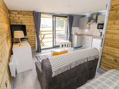 A living area with a sofa and throw blanket facing a television on a white cabinet next to sliding glass doors with curtains and a view of a hot tub outside a kitchen with cabinets and appliances at Cilan Lodge in Llandrillo