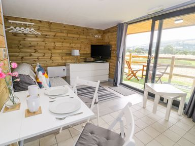 A small living room with a dining table set for two a white dresser with a TV and a sliding glass door leading to an outdoor seating area at Cilan Lodge in Llandrillo