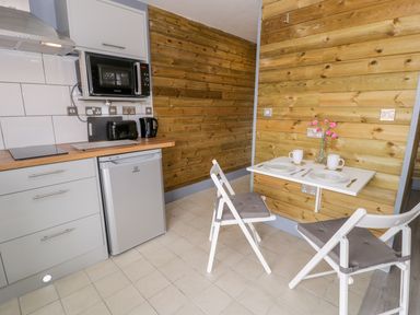 A small kitchen area with white cabinets a microwave a toaster and a kettle next to a fold-down table with two place settings and chairs at Cilan Lodge in Llandrillo