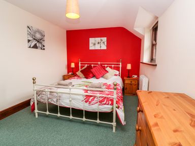 A bedroom with a double bed with floral duvet and cushions two wooden bedside tables with lamps a wooden chest of drawers and a window at Mossley Cottage in St Buryan