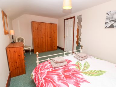 A bedroom with a bed featuring floral bedding a wooden wardrobe a wooden chest of drawers a chair a lamp and a door at Mossley Cottage in St Buryan