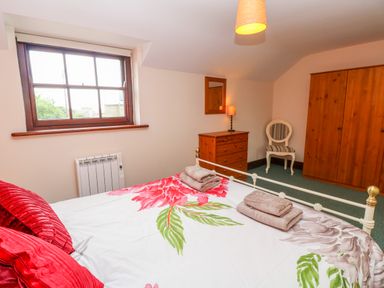 A bedroom with a floral bedspread red pillows a wooden dresser a striped chair a wooden wardrobe and a window at Mossley Cottage in St Buryan
