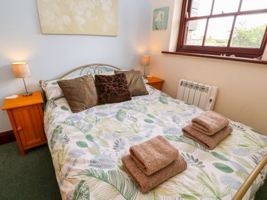 A bedroom with a bed with leaf-patterned bedding pillows bedside tables with lamps and folded towels on Mossley Cottage in St Buryan