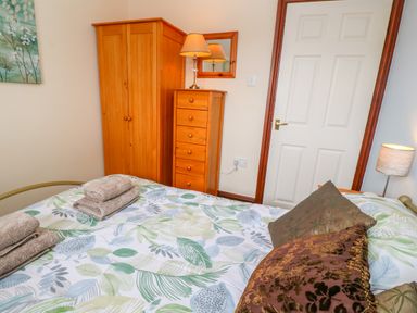 A bedroom with a bed covered in leaf-patterned bedding two brown pillows a wooden wardrobe and chest of drawers with a lamp on top a door and a wall lamp at Mossley Cottage in St Buryan