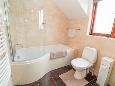 A bathroom with a bathtub and shower, a toilet, a towel rack with a towel, a radiator, and a small cabinet under a window at Mossley Cottage in St Buryan