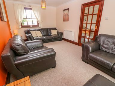 A living room with three dark leather sofas a window with white curtains a wooden door with glass panels and a lamp at Mossley Cottage in St Buryan