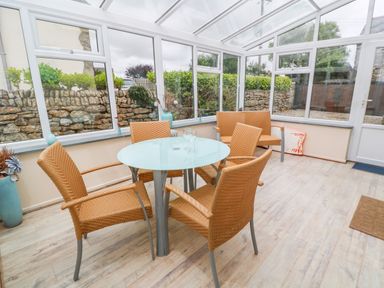 A conservatory with a round table and wicker chairs overlooking a stone wall garden at Mossley Cottage in St Buryan