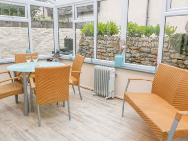 A conservatory with a round glass table four wicker chairs a wicker bench a heater and vases with flowers at Mossley Cottage in St Buryan