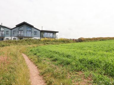 A pathway leading to modern houses with glass balconies next to a green field at Sandpiper in Perranporth