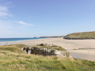 A sandy beach with grassy cliffs and blue ocean under a clear sky at Sandpiper in Perranporth