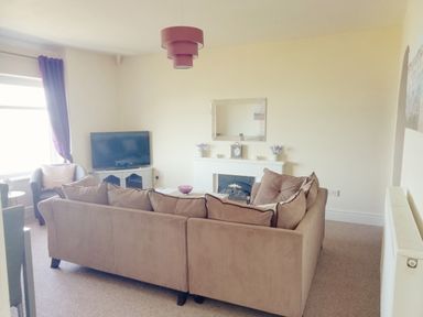 A living room with a beige sofa, a TV on a white stand, a fireplace with a clock and flowers, and a purple lampshade at Llys Madoc The Top Flat in Penmaenmawr