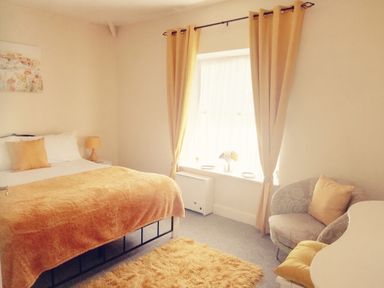 A bedroom with a bed yellow blanket and pillow a bedside table lamp a window with yellow curtains a chair with a yellow cushion and a rug at Llys Madoc The Top Flat in Penmaenmawr