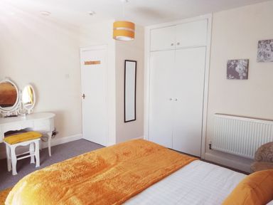 A bedroom with a bed covered in an orange blanket a white dressing table with mirrors an orange cushioned stool a wall-mounted mirror closed white closet doors radiator and wall art at Llys Madoc The Top Flat in Penmaenmawr