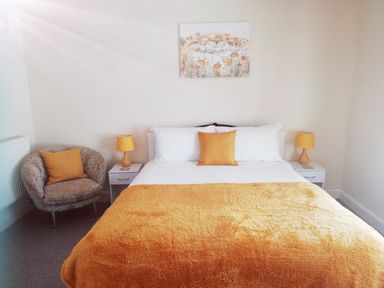 A bedroom with a bed with white and orange bedding two bedside tables with lamps a chair with an orange cushion and a painting above the bed at Llys Madoc The Top Flat in Penmaenmawr