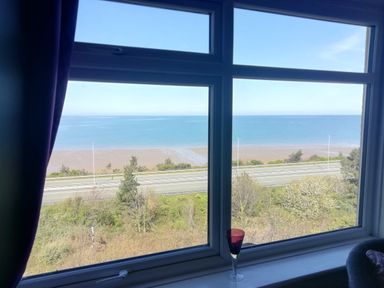 A window view showing a road, trees, a beach, and the sea at Llys Madoc The Top Flat in Penmaenmawr