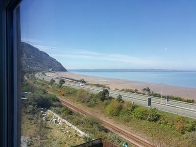 A coastal view with a mountain, roads, railway tracks, and a beach at Llys Madoc The Top Flat in Penmaenmawr